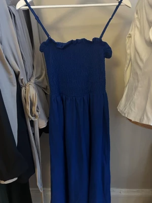 Marinblå långklänning med smock - Säljer en marinblå långklänning med smala axelband och smockad överdel. Klänningen har en lätt rynkad volang upptill och faller rakt ner i en luftig modell. Perfekt för varma dagar och enkel att styla med accessoarer.