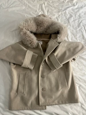 Beige kappa  från Gina Tricot - Säljer en beige kappa från Gina Tricot i storlek M. Jackan har en stor huva med mjuk fuskpäls, raka ärmar med uppvik, knäppning med fem knappar framtill och två fickor. Perfekt för kalla dagar och ger en clean och stilren look.