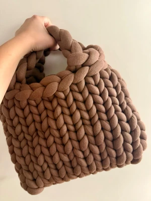 Brun chunky handgjord handväska 🤍 - Unik handväska i chunky stickad design med breda flätor i mjukt brunt material. Väskan har ett rejält handtag och en cool, oversized look som verkligen sticker ut. Perfekt för dig som vill ha något annorlunda och trendigt till din outfit.