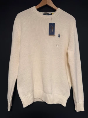 Vit stickad tröja från Polo Ralph Lauren - Klassisk vit stickad tröja från Polo Ralph Lauren med rund halsringning och marinblå broderad logga på bröstet. Tröjan har ribbade muddar vid ärmslut och nederkant, perfekt för en clean och stilren look.