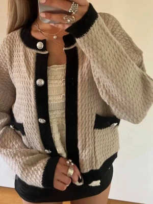 Beige kofta med svarta detaljer - Säljer en beige kofta med svarta kontrastkanter och stora dekorativa knappar. Koftan har ett stickat mönster, två fickor framtill och långärmad passform. Perfekt för dig som gillar en chic och trendig look.