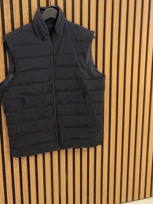 Svart dunväst från MNG Essentials - Säljer en svart dunväst från MNG Essentials med hög krage och två dragkedjeförsedda fickor framtill. Västen har quiltad design och stängs med dragkedja. Perfekt för lager-på-lager och enkel att matcha till olika outfits.