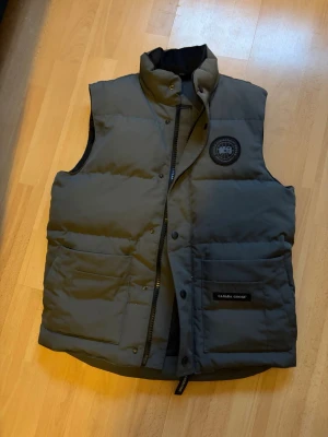 dunväst från Canada Goose - Säljer en grå dunväst från Canada Goose med hög krage, två stora fickor framtill och diskret logga på bröstet. Västen har dragkedja och tryckknappar samt en stilren, quiltad design. Perfekt för lager-på-lager under kyliga dagar.