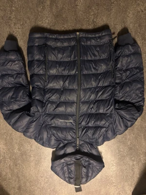 Mörkblå dunjacka Peak Performance S - Säljer en mörkblå dunjacka från Peak Performance i storlek S. Jackan har huva, dragkedja framtill och quiltad design. Perfekt för kalla dagar och riktigt skön att bära. Klassisk look med Peak Performance-logga på bröstet.