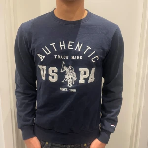 Marinblå sweatshirt från U.S. Polo Assn - Säljer en marinblå sweatshirt från U.S. Polo Assn med stort tryck framtill och klassisk rund hals. Tröjan har långa ärmar och ribbade muddar vid ärmslut och nederkant. Perfekt för dig som gillar sportig och avslappnad stil.