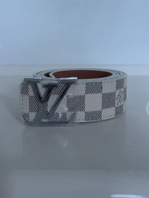 Rutig Louis Vuitton bälte - Säljer ett snyggt bälte från Louis Vuitton med klassiskt rutigt mönster i vitt och grått. Bältet har en stor LV-logga i silver som spänne och insidan är brun. Perfekt accessoar för att lyfta vilken outfit som helst. Kvitto ingår 