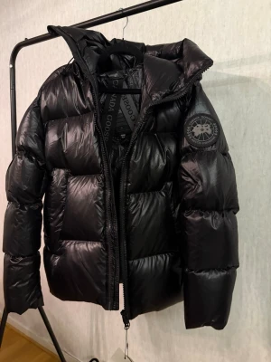 Svart pufferjacka från Canada Goose - Säljer en svart pufferjacka från Canada Goose i storlek S. Jackan har en hög krage, stora vadderade partier och dragkedja framtill. Märkets logga sitter på ärmen och insidan har praktiska axelband för att bära jackan som en ryggsäck. Perfekt för kalla dagar.