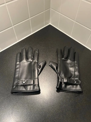 Svarta handskar knappdetalj - Snygga svarta handskar med stilren design. Handskarna har en dekorativ knappdetalj vid handleden och slät yta som ger en clean look. Perfekta för dig som vill ha ett klassiskt och enkelt accessoar till höst och vinter. Passar aveage man/storelk 9 