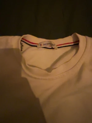 Vit sweatshirt från Moncler - Säljer en stilren vit sweatshirt från Moncler med rund hals och klassisk logga broderad på bröstet. Tröjan har långa ärmar och ribbade muddar. Insidan av kragen har en snygg randig detalj i rött, vitt och blått. Perfekt för dig som gillar clean och exklusiv stil.