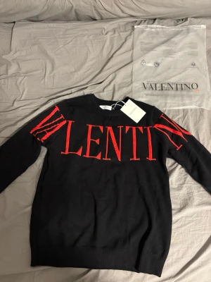 Svart tröja från Valentino med röd logga - Säljer en svart tröja från Valentino med stor röd logotyp över bröstet och axlarna. Tröjan är i 100% bomull och har rund hals samt långa ärmar. Perfekt för dig som vill ha en statement piece i garderoben.