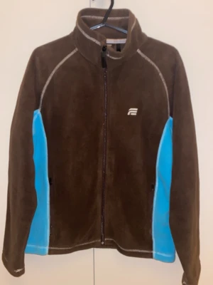 Brun fleecejacka Everest med blå detaljer - Mysig brun fleecejacka från Everest i storlek 36. Jackan har ljusblå paneler på sidorna, hel dragkedja framtill och hög krage. Perfekt för kyliga dagar och outdoor-häng. Broderad Everest-logga på bröstet och nacken.