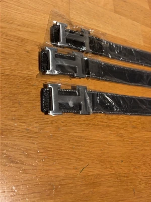 Svart bälte med H-spänne. 185kr styck - Säljer ett svart bälte med ett stort H-format metallspänne. Bältet är gjort i präglat skinnliknande material och har vita sömmar längs kanterna. Perfekt accessoar för att lyfta din outfit. 