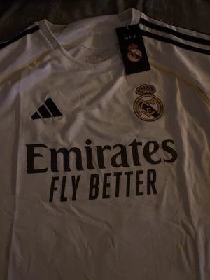 Real Madrid match ställ - Säljer en officiell Real Madrid match ställ i storlek L. Tröjan är vit med svarta och gula detaljer, Real Madrids klubbmärke på bröstet och Adidas-logga. Tillverkad i lätt och ventilerande polyester, perfekt för fotboll eller som supporterplagg.