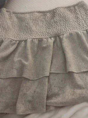 Beige volangkjol med smock - Supersöt beige grön  kjol med bred smock i midjan och dubbla volanger. Kjolen har en mjuk struktur och känns riktigt flowy och bekväm. Perfekt för dig som gillar en enkel men ändå trendig look.