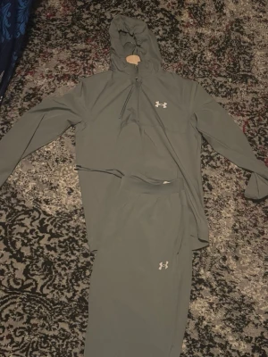 Under armour tracksuit - Snygg och bekväm Under Armour tracksuit i stilren grön/grå färg. Setet består av en hoodie med halv dragkedja och huva samt matchande byxor. Tillverkad i ett lätt och stretchigt material som är perfekt för träning, promenader eller vardagsbruk. Diskret Under Armour-logga på både tröja och byxor ger en sportig och clean look.