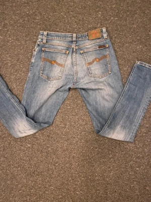 Nudie Jeans blå skinny jeans W29 L30 - Säljer ett par ljusblå skinny jeans från Nudie Jeans med klassisk orange söm och broderad detalj på bakfickorna. Modellen har normal passform och är tillverkad i mjukt jeansmaterial. Perfekt för dig som gillar stilrena och bekväma jeans.