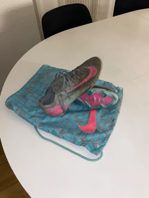 Nike fotbollsskor turkos /rosa - Säljer ett par Nike fotbollsskor i turkos med rosa detaljer och logga. Skorna har snörning, låg profil och en sula med rosa och gröna inslag. Ovandelen är i syntetmaterial och innersulan är röd. Medföljer en blå Nike-påse med tryck.