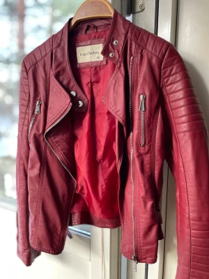 Vinröd jacka från King of Fashion - Säljer en vinröd jacka från King of Fashion i storlek XS. Jackan har coola detaljer som dragkedjor, knappar och quiltade partier på axlarna. Snygg bikerstil med asymmetrisk dragkedja och flera fickor framtill. Perfekt 