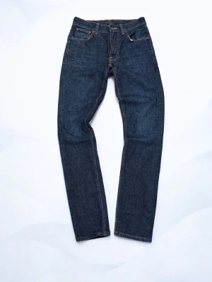 Nudie Jeans - Säljer ett par mörkblå jeans från Nudie Jeans med klassisk femficksdesign och kontrastsömmar. Modellen har en tight passform och raka ben. Tillverkade i slitstarkt jeansmaterial av bomull. Snygg läderpatch bak i midjan med Nudie-logga.