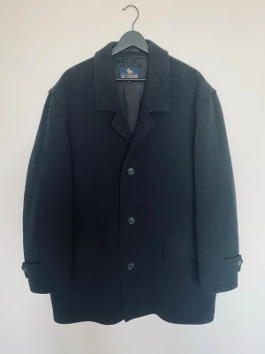 Svart rock från MC Gordon XL - Klassisk svart rock från MC Gordon i storlek XL. Den har en rak passform. Materialet är 70% ull och 30% polyester. Perfekt för kyliga dagar och har snygga detaljer vid ärmsluten.