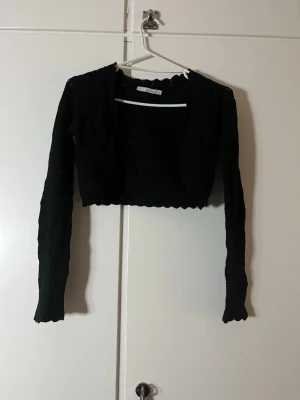 Svart bolerotopp från More & More - Kort svart bolerotopp från More & More i virkad look med vågig kant och lång ärm. Toppen är genombruten och har ett elegant mönster i tyget som ger en snygg struktur. Perfekt att slänga över ett linne eller kl soppa till en outfit.