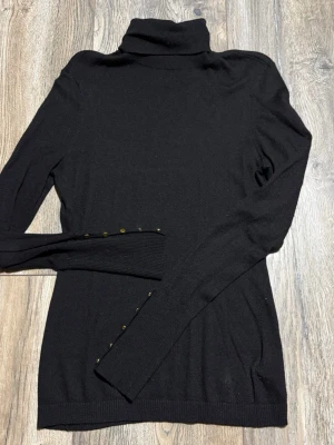 Svart polotröja från H&M med knappar - Säljer en svart polotröja från H&M i storlek S. Tröjan har långa ärmar och snygga guldfärgade knappar längs ärmsluten. Mjuk och tunn stickad kvalitet som passar perfekt till lager på lager under höst och vinter.