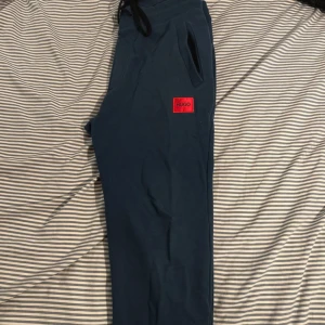 Blå mjukisbyxor från Hugo - Snygga blå mjukisbyxor från Hugo med svart snörning i midjan och röd logotyp-patch på benet. Byxorna har två sidofickor och en bakficka, samt elastisk midja för skön passform. Perfekta för chill eller träning.