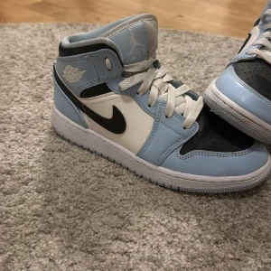 Nike Air Jordan 1 Mid UNC blå/vit - Nike Air Jordan 1 Mid sneakers i UNC färgway med ljusblå, vit och svart skinnpanel. Klassisk Jordan-logga på sidan, svart swoosh och perforerad svart tå. Skon har snörning och en platt sula, perfekt för dig som gillar streetstyle och ikoniska sneakers.