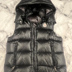 Svart dunväst från Moncler  - Svart dunväst från Moncler med avtagbar huva och två dragkedjeförsedda fickor. Västen har quiltad design och Moncler-logga på bröstet. Materialet är glansigt och känns lyxigt, perfekt för att hålla värmen under kyliga dagar. Stängs med dragkedja framtill.