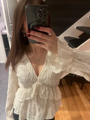 Vit omlottblus med volanger - Säljer en vit blus med omlott och v-ringad design. Blusen har långa ärmar med volangkant och är tillverkad i ett krinklat, lätt material. Använd fåtal gånger och är i bra skick.