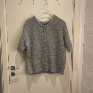 Grå kofta från Gina Tricot - Mysig grå kofta från Gina Tricot i storlek L. Koftan har korta ärmar, rund hals och knappar framtill. Materialet är fluffigt och stickat, perfekt för att ge en chill vibe till din outfit. Enkel att matcha med jeans eller kjol.