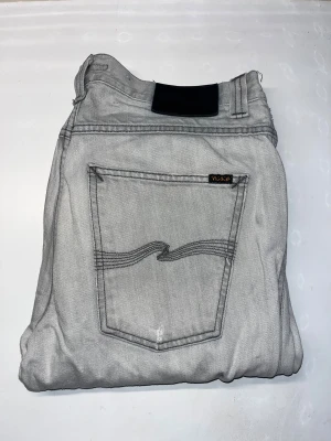 Nudie Jeans  - Säljer ett par ljusgrå Nudie Jeans Slim Jim. Jeansen har en slim passform. Materialet är mjukt jeans-tyg med svarta sömmar. Midja 42cm, längd 109cm. JAG SKICKAR EJ FLER BILDER NÄR JAG BÄR JEANSEN! för storleksguide hänvisar jag till måtten och för bättre inblick i passformen rekommenderar jag att googla på modellnamnet. kolla måtten noggrant :) OBS se defekterna på de 2 sista bilderna. S3