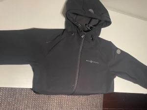 Svart vindjacka från Sail Racing storlek XS - Svart vindjacka från Sail Racing med huva och snygga logodetaljer på bröstet och ärmen. Jackan har dragkedja framtill och justerbar huva med snörning. Perfekt för dig som gillar stilren och sportig look.
