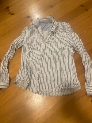 Randig skjorta från Hampton Republic - Snygg vit skjorta med blåa vertikala ränder från Hampton Republic. Klassisk krage, långärmad och med knappar framtill. Skjortan är gjord i mjuk bomull och har en stilren look som funkar till jeans eller chinos. Perfekt för dig som gillar preppy stil.