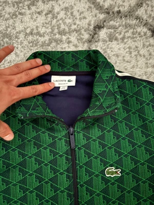 Grön mönstrad trackjacka Lacoste - Snygg grön trackjacka från Lacoste med geometriskt mönster och klassisk krokodillogga på bröstet. Jackan har svarta muddar, hel dragkedja och vita ränder längs ärmarna. Perfekt för dig som gillar sportig och modern stil.