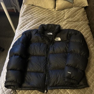 Svart pufferjacka från The North Face - Säljer en svart pufferjacka från The North Face, modell 700. Jackan har hög krage, dragkedja framtill och logga på bröstet och ärmen. Perfekt för kalla dagar med sin fluffiga design. Lite lagad men syns knappt