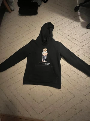 Mörkblå Polo Bear hoodie från Ralph Lauren - Mörkblå hoodie från Ralph Lauren med ikoniskt Polo Bear-motiv på bröstet. Tröjan har huva med snörning, långärmad design och en stor magficka. Materialet är mjuk bomull och polyesterblandning, Snyggt tryck med björnen i solglasögon och randig tröja. Använd ett fåtal gånger men ser ut som helt ny. Inga defekter! Fraktar på 1-2 dagar! 