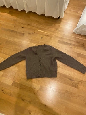 Brun ribbad kofta med knappar - Mysig brun kofta med ribbad struktur och fyra knappar framtill. Modellen har v-ringad hals och långa ärmar med breda muddar. Perfekt för lager-på-lager. Materialet känns stickat och är skön. Lapp saknas men den är i en XS.