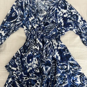 Blåvit mönstrad långärmad klänning - Säljer en blåvit klänning med coolt abstrakt mönster och v-ringad hals. Klänningen har lång ärm, smock i midjan och volangdetaljer som ger en snygg siluett. Perfekt för dig som gillar att sticka ut med färg och mönster.