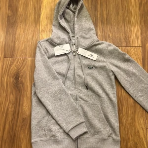 Grå hoodie från Lacoste - Säljer en stilren grå hoodie från Lacoste med dragkedja . Perfekt för en avslappnad look. Sitter som en S men står M i koftan, helt ny.