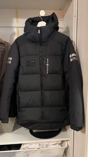 Svart Sail Racing dunjacka S - Svart dunjacka från Sail Racing med huva, dragkedja och coola loggor på ärmar och bröst. Jackan är vind- och vattenavvisande, andas och har certifierat dun. Perfekt för kalla dagar och har en clean, sportig look. Har en liten skada vid ena fickan som inte syns så mycket. Köp för 6000 knappt använd en hel vinter.
