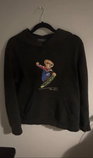Svart hoodie Polo Bear Ralph Lauren - Svart hoodie från Polo Ralph Lauren med coolt Polo Bear-tryck på bröstet där björnen åker . Klassisk huva och stor magficka framtill. Trycket har färger som blått, rött och grönt. Perfekt för dig som gillar streetwear och vill sticka ut.