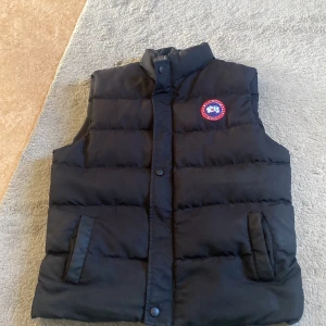 Svart dunväst från Canada Goose - Säljer en svart dunväst från Canada Goose med klassisk logotyp på bröstet. Västen har ståkrage, knappar och två fickor framtill. Materialet består av polyester och polyuretan, det  gör den både vindtät och vattentät. Västen är i ett mycket bra skick dock har den  ett hål i högra fickan.  västen passar perfekt till dom kyliga vår/sommar kvällarna och är en fantastisk  väst. Det går  att byta mot något stiligt Endast väst vinterjacka och  cardigan. Man kan inte lämna tillbaka efter ja har fraktat 