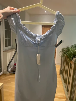 Ljusblå kort klänning från NLY Trend - Supersöt ljusblå klänning från NLY Trend med kort längd och slim fit. Klänningen har offshoulder-ärmar, volangdetaljer runt halsringningen och en knytning framtill. Perfekt för dig som gillar romantisk och feminin stil. Materialet känns mjukt och lätt mot huden.