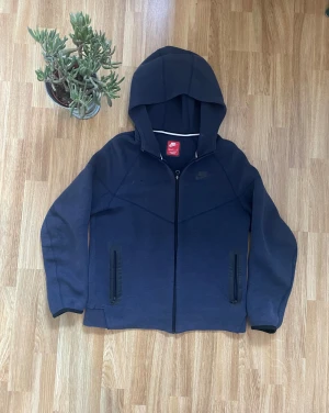 Nike Tech Fleece zip hoodie blå/niebieskie buty nike tech - Snygg mörkblå Nike Tech zip-hoodie med dragkedja och huva. Bekväm, lätt och värmande – perfekt till vardag eller streetwear. ✔️ Fickor med dragkedja ✔️ Slim fit, klassisk Tech-design  Storlek: 158/170 Skick: 8/10, har ett litet håll som syns vid bilderna  Nypris: 1200 kr.                            Ser smutsig ut då jag ej har tvättat den men tvättar alltid innan frakt 
