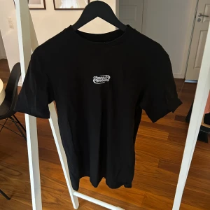 Svart t-shirt från FSBN, relaxed fit - Svart t-shirt från FSBN i relaxed fit, storlek S. T-shirten har korta ärmar och ett litet vitt tryck med texten 'chaotic system' på bröstet. Tillverkad i mjuk bomull, perfekt för en chill och enkel stil.