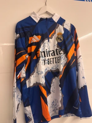 Real Madrid långärmad träningströja Adidas - Säljer en långärmad Real Madrid träningströja från Adidas med dragkedja vid halsen. Tröjan har ett grafiskt mönster i blått, vitt, orange och grått, samt klubbmärke och Adidas-logga på bröstet. Materialet är lätt och andas, perfekt för fotbollsträning.