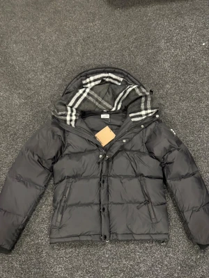 Svart pufferjacka från Burberry - Svart pufferjacka från Burberry med klassisk rutig huva i grått och vitt. Jackan har dragkedja och knappar framtill, två sidofickor med dragkedja och en snygg, quiltad design. Perfekt för kalla dagar med sin tjocka och fluffiga look samt som julklapp!