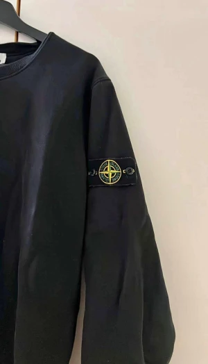 Svart sweatshirt från Stone Island - Svart långärmad sweatshirt från Stone Island med klassisk rund hals och ikonisk patch med logga på vänster ärm. Tröjan är tillverkad i mjuk bomull och har en clean, normal passform som funkar till jeans eller cargo. Perfekt för dig som gillar streetwear och snygga detaljer.