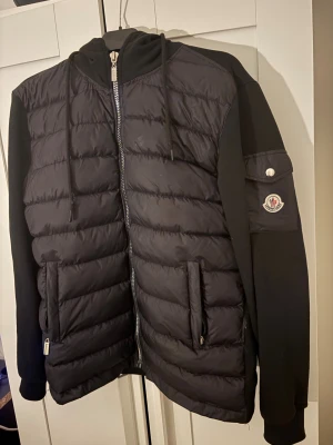 Svart pufferjacka Moncler L - Svart pufferjacka från Moncler i storlek L Jackan har quiltad framsida, huva med snörning, dragkedja och två fickor med dragkedja. Ärmarna är släta med Moncler-logga på vänster arm. Perfekt för dig som gillar stilren och sportig look.